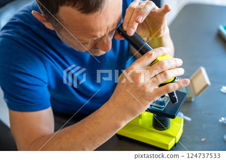 Man examining various samples under a microscope at home as a hobby. Concept of scientific curiosity and amateur microscopy Man examining various samples under a microscope at home as a hobby. Concept of scientific curiosity and amateur microscopy 124737533