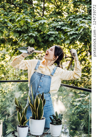 Young woman joyfully tending to potted plants on a balcony surrounded by lush trees. Emotional support plants, nurturing routines, Gen Z hobbies, sustainable living 124739772
