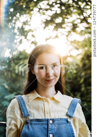Young woman with brown hair standing calmly outdoors in front of trees and warm sunlight. Self-love, emotional wellness, authentic happiness, personal empowerment 124739777