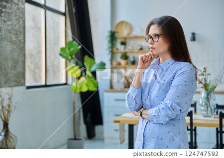 Portrait of serious middle aged woman looking out window in home Portrait of serious middle aged woman looking out window in home 124747592