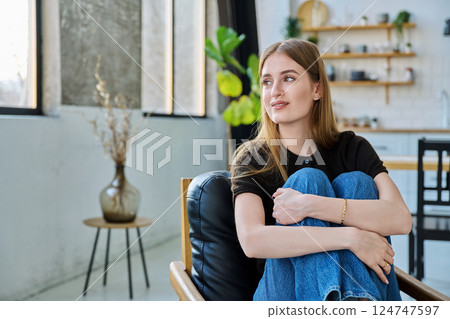 Portrait of young beautiful smiling woman looking at window sitting at home in chair 124747597
