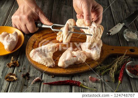 A man skillfully threads raw chicken wings onto a metal fork, preparing them for cooking. Surrounding ingredients include herbs, spices and garlic on a rustic wooden table A man skillfully threads raw chicken wings onto a metal fork, preparing them for cooking. Surrounding ingredients include herbs, spices and garlic on a rustic wooden table 124748707