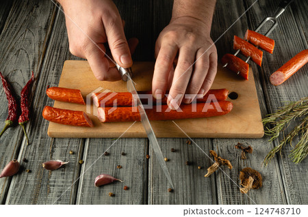 Hands expertly slice spicy sausages on a wooden cutting board, surrounded by herbs and spices. The warm, rustic kitchen atmosphere enhances the culinary experience 124748710