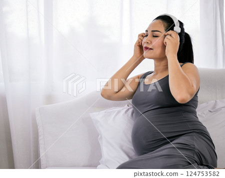 Close-up of pregnant woman in gray dress sitting on bed with eyes closed enjoying music through headphones in bedroom with copy space Close-up of pregnant woman in gray dress sitting on bed with eyes closed enjoying music through headphones in bedroom with copy space 124753582