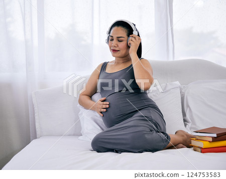 Pregnant woman in gray dress relaxing on bed with one hand on belly and listening to music with headphones beside stack of colorful books 124753583