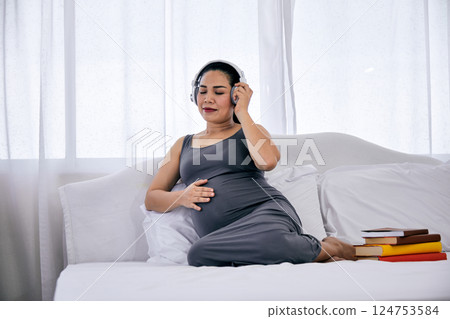 Pregnant woman in gray dress lying on bed while touching belly and listening to music with headphones next to stack of colorful books in bedroom 124753584