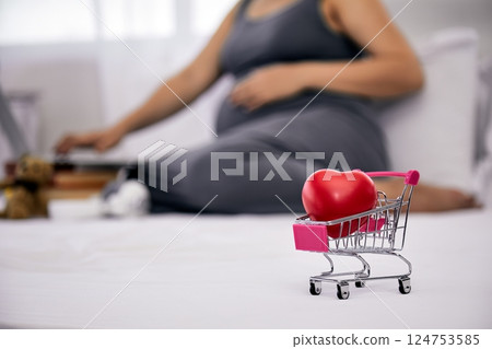 Mini shopping cart with red heart in focus on bed with pregnant woman in gray dress using laptop in background; love care online shopping and motherhood 124753585