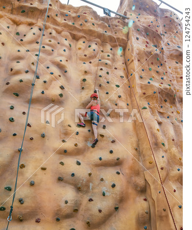 Young climber ascends a rock wall during a summer day at an outdoor adventure park in a vibrant setting 124754243