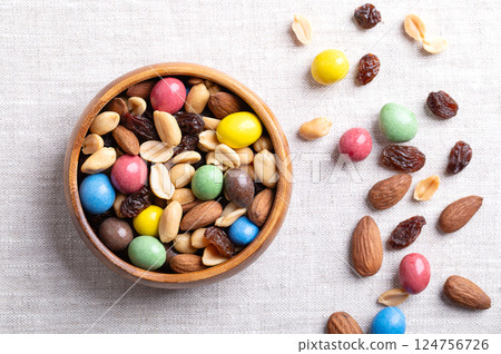 Rainbow trail mix in a wooden bowl on linen fabric. A snack mix of candy coated milk chocolate peanuts, roasted peanuts, raisins, and roasted almonds. A combination of sweets, dried fruits, and nuts. 124756726