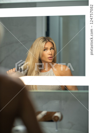 In chic bathroom woman is using round brush style her long hair while gazing her reflection in well lit mirror. Soft colors create calming atmosphere. 124757082