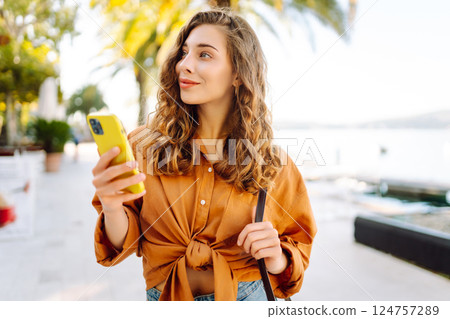 Portrait of a young woman holding a phone in her hands on a city avenue, on a sunny day. 124757289
