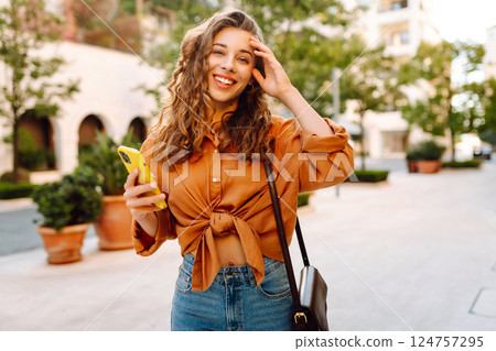 Portrait of a young woman holding a phone in her hands on a city avenue, on a sunny day. 124757295