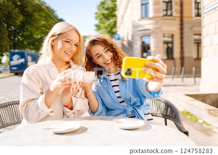 Young women taking selfie while sitting on terrace on sunny day. Girlfriends drink coffee with phone Young women taking selfie while sitting on terrace on sunny day. Girlfriends drink coffee with phone 124758283