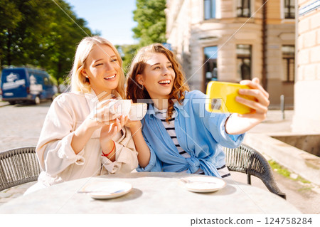 Young women taking selfie while sitting on terrace on sunny day. Girlfriends drink coffee with phone Young women taking selfie while sitting on terrace on sunny day. Girlfriends drink coffee with phone 124758284