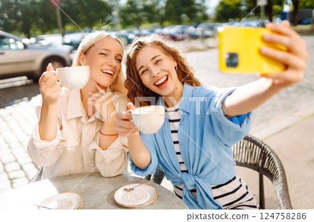 Young women taking selfie while sitting on terrace on sunny day. Girlfriends drink coffee with phone Young women taking selfie while sitting on terrace on sunny day. Girlfriends drink coffee with phone 124758286