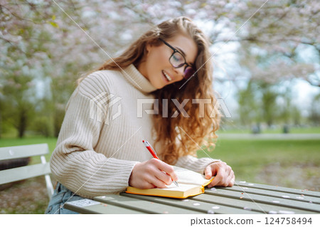 Young woman sitting at a table in blooming spring park with a notepad and laptop. Lifestyle concept. Young woman sitting at a table in blooming spring park with a notepad and laptop. Lifestyle concept. 124758494