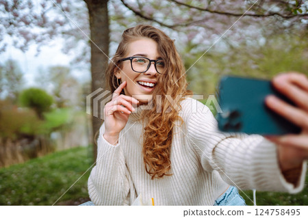 Young woman takes selfie against background of flowering trees in park. Concept of relaxation, fun. 124758495