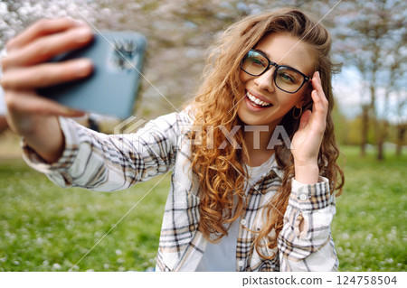 Young woman takes selfie against background of flowering trees in park. Concept of relaxation, fun. Young woman takes selfie against background of flowering trees in park. Concept of relaxation, fun. 124758504
