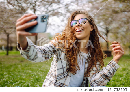 Young woman takes selfie against background of flowering trees in park. Concept of relaxation, fun. 124758505