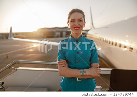 A smiling flight attendant standing at sunset beside an airplane, radiating warmth and joy A smiling flight attendant standing at sunset beside an airplane, radiating warmth and joy 124759744