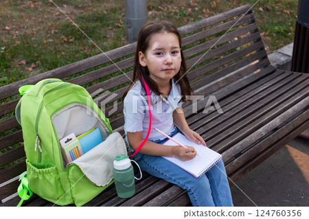 Young girl drawing on bench with backpack and school supplies in park 124760356