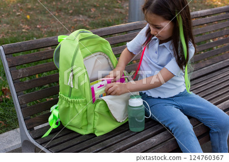 Young girl organizing backpack on park bench 124760367