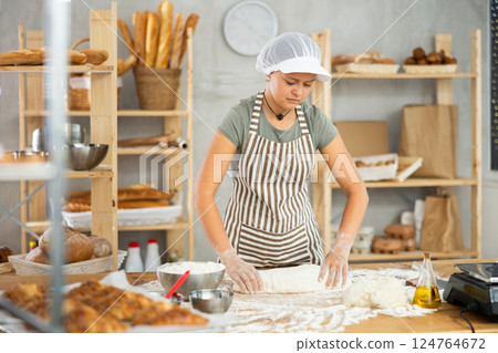 Female baker prepares raw dough in bakery, forming it into long sausages to be cut into pieces for baking buns or croissants 124764672