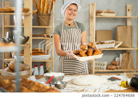 Smiling female baker in uniform stands behind counter in small private bakery offering croissants 124764686