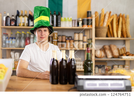 Smiling man seller in traditional Irish green cap offers Irish ale on St. Patrick Day in supermarket 124764751