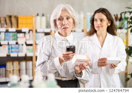 Senior woman in pharmacy scans QR code on omega 3 supplement, woman pharmacist in background 124764794