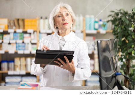 Mature female pharmacist makes notes on electronic tablet - keeps track of receipt and expenditure of pharmacy Mature female pharmacist makes notes on electronic tablet - keeps track of receipt and expenditure of pharmacy 124764811