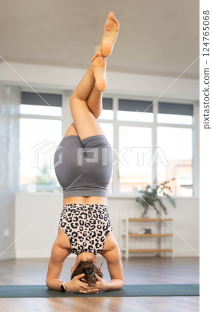 Young girl hatha yoga coach tries to perform various variations of feathered peacock asana, pincha mayurasana in gym hall 124765068