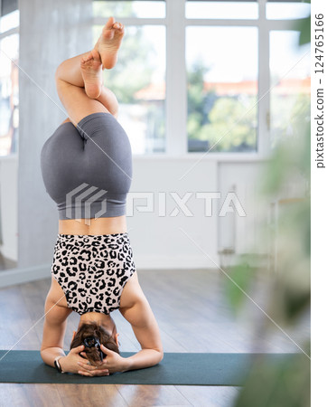 Experienced sporty young girl fitness club teacher performs feathered peacock asana, pincha mayurasana in fitness room 124765166