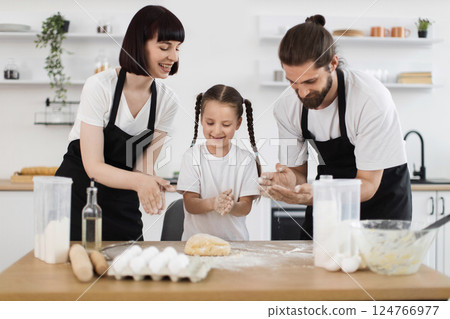 Smiling young parents and daughter in kitchen making dough, sharing happy bonding moment together Smiling young parents and daughter in kitchen making dough, sharing happy bonding moment together 124766977