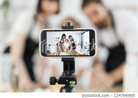 Caucasian family consisting man, woman, child recording cooking tutorial while kneading dough. Scene captures modern lifestyle with teamwork in home setting. 124766980