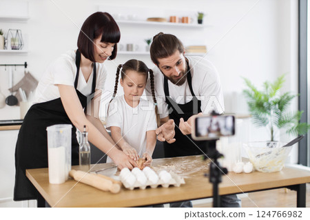 Caucasian parents and daughter cooking together in bright kitchen, wearing aprons, smiling happily Caucasian parents and daughter cooking together in bright kitchen, wearing aprons, smiling happily 124766982