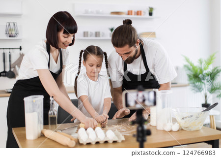Caucasian family of three, including young girl and two adults, kneading dough on table while creating cooking vlog in modern kitchen environment emphasizing family connection, creativity, Caucasian family of three, including young girl and two adults, kneading dough on table while creating cooking vlog in modern kitchen environment emphasizing family connection, creativity, 124766983