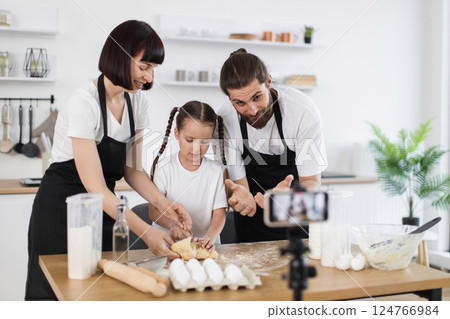 Caucasian family including parents and child preparing dough in kitchen and recording themselves, engaging in cooking activity promoting fun and togetherness while sharing their skills. Caucasian family including parents and child preparing dough in kitchen and recording themselves, engaging in cooking activity promoting fun and togetherness while sharing their skills. 124766984
