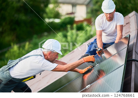 Workers building photovoltaic solar panel system on rooftop of house. Men technicians in helmets and gloves installing solar module with help of hex key outdoors. Renewable energy generation concept. 124768661