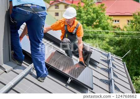 Workers building solar panel system on rooftop of house. Two men installers in helmets installing photovoltaic solar module outdoors. Alternative, green and renewable energy generation concept. Workers building solar panel system on rooftop of house. Two men installers in helmets installing photovoltaic solar module outdoors. Alternative, green and renewable energy generation concept. 124768662