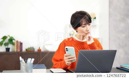 A middle-aged woman worried while looking at her smartphone and laptop A middle-aged woman worried while looking at her smartphone and laptop 124768722