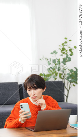 A middle-aged woman worried while looking at her smartphone and laptop 124768779