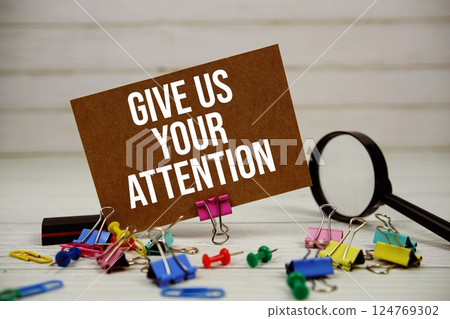 Give Us Your Attention text on paper card with magnifying glass and stationary on wooden background 124769302