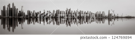 Monochrome city skyline with tall skyscrapers reflecting on calm water under a cloudy sky, Dubai Marina bay UAE 124769894