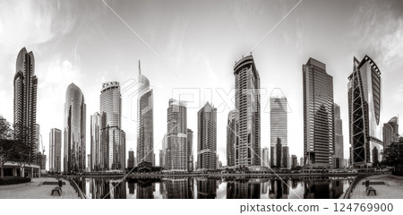 Monochrome city skyline with tall skyscrapers reflecting on calm water under a cloudy sky, Dubai Marina bay UAE 124769900