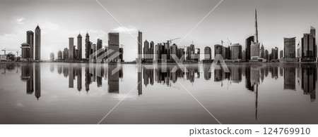 Monochrome city skyline with tall skyscrapers reflecting on calm water under a cloudy sky, Dubai Marina bay UAE 124769910