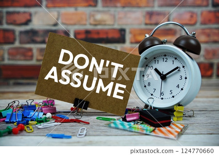 Don't Assume text on paper card with alarm clock and stationary on wooden background 124770660