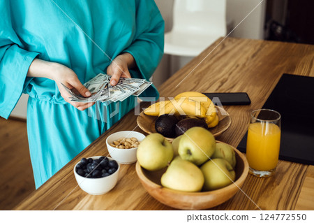 Close-up of woman counting US dollar bills beside a kitchen counter with fruit and nuts. Health is wealth, lifestyle investment, food budgeting, mindful living Close-up of woman counting US dollar bills beside a kitchen counter with fruit and nuts. Health is wealth, lifestyle investment, food budgeting, mindful living 124772550