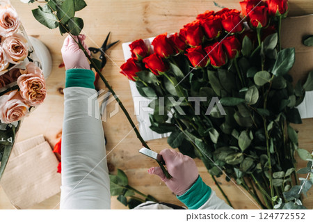 Florist trimming red rose stems by hand with a knife beside fresh bouquets and greenery. Bouquet assembly, floristry craftsmanship, slow production, floral detailing, hands-on work Florist trimming red rose stems by hand with a knife beside fresh bouquets and greenery. Bouquet assembly, floristry craftsmanship, slow production, floral detailing, hands-on work 124772552