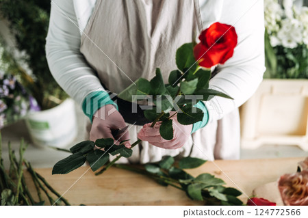 Florist wearing gloves trims red rose stems at a wooden worktable in a flower studio. Revival of local floral businesses, shop small, ethical flowers, community-based commerce 124772566